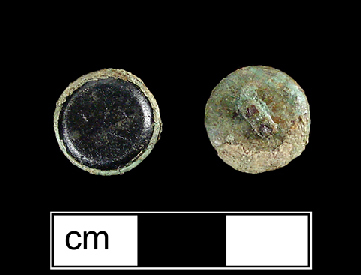 Copper alloy button back with a dark black or purple glass inset, flat on the front and beveled at the edge. The copper corrosion of the button back is toxic to microorganisms, which likely explains how some fabric that was in contact with the button survived and is still attached to the button back. The weave is visible, and the fabric is probably linen.